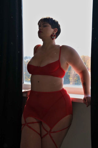 Комплект KLEO червоного кольору – бюстгальтер, високі трусики та декоративні гартери, модель стоїть біля вікна, м’яке природне світло