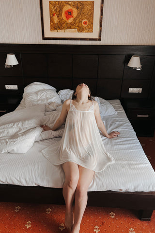 Жіноча напівпрозора сорочка Vanda молочного кольору з легкої сітки, вигляд спереду, модель сидить на ліжку в інтер’єрі спальні, м’який силует і повітряна посадка.